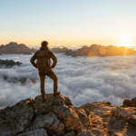 Eine Person steht siegreich auf einem Berggipfel über dem Nebel bei Sonnenaufgang als Symbol für die 5 Schritte aus dem Tief.